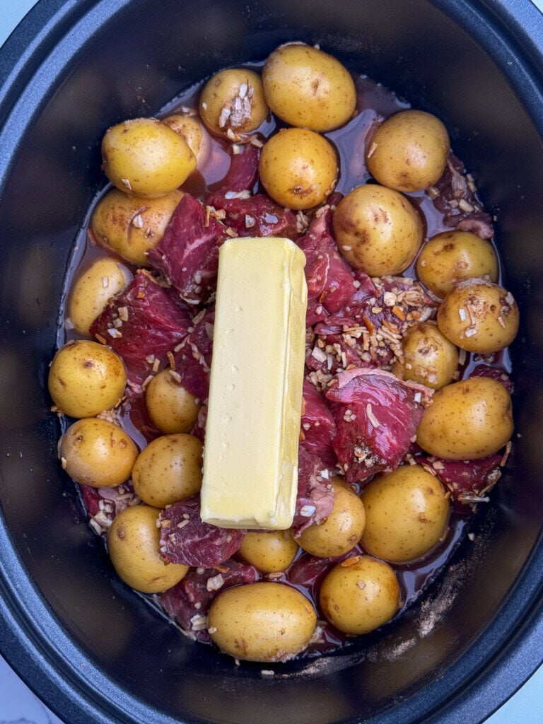 A slow cooker filled with whole baby potatoes, chunks of beef, seasoning, and a whole stick of butter placed on top, ready to be cooked.
