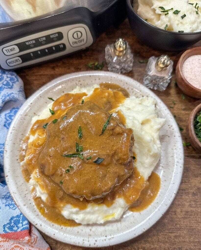 A plate of mashed potatoes topped with a round piece of meat covered in brown gravy, garnished with chopped herbs—a perfect Cooking Challenge dish. Nearby are bowls of mashed potatoes, parsley, a slow cooker, salt and pepper shakers, and a blue napkin.
