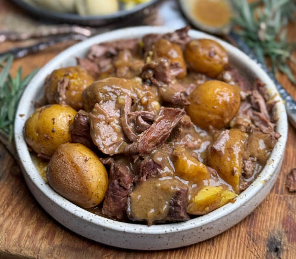 A bowl of pot roast with chunks of tender beef, golden potatoes, and rich brown gravy, served on a wooden table with sprigs of fresh rosemary nearby.