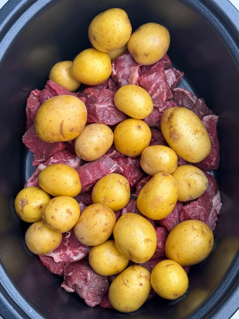 Raw yellow potatoes are piled on top of chunks of raw beef in a black slow cooker, ready to be cooked.
