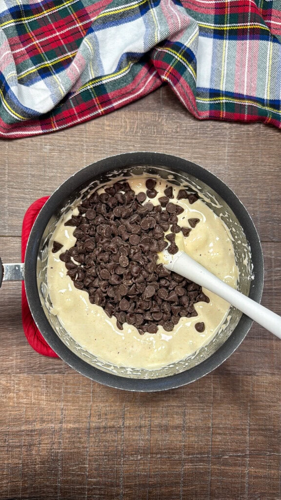 A mixing bowl with thick batter and a pile of chocolate chips on top, being stirred with a white spatula. A plaid cloth is visible on the wooden surface in the background.