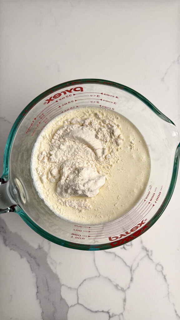 A glass measuring cup containing milk and a mound of flour, set on a white marble countertop.