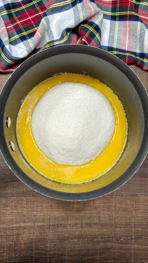 A saucepan containing melted butter with a mound of granulated sugar in the center, placed on a wooden surface with a colorful plaid cloth in the background.