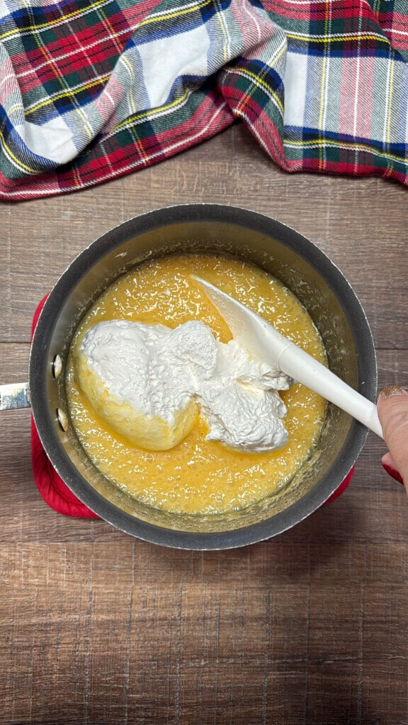 A saucepan containing a melted butter with sugar. There is a pile of marshmallow cream in the saucepan and a white spatula. A plaid cloth is nearby on a wooden surface.