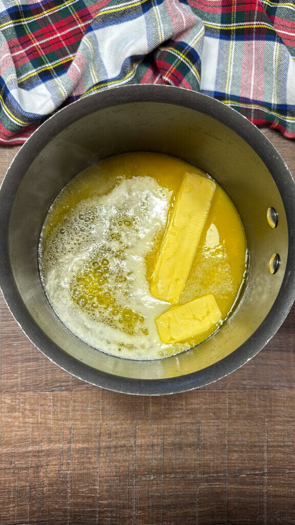 A saucepan with melting butter sticks, some already liquid, sits on a wooden surface. A colorful plaid cloth is partially visible at the top of the image.