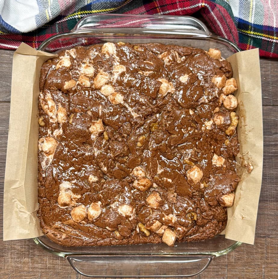 A glass baking dish filled with a chocolate marshmallow fudge sits on a wooden surface, lined with parchment paper, with a colorful plaid cloth nearby.