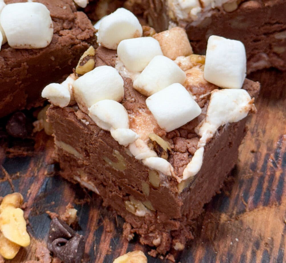 A close-up of a piece of chocolate fudge topped with mini marshmallows and chopped walnuts, resting on a wooden surface with more nuts and chocolate chips scattered around.