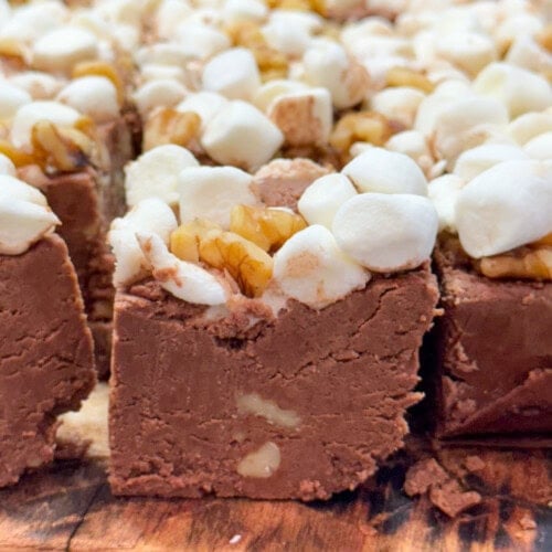Close-up of chocolate fudge squares topped with mini marshmallows and walnut pieces, arranged on a wooden surface.
