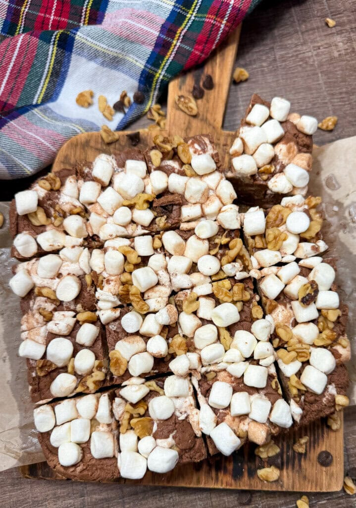 A batch of chocolate fudge topped with mini marshmallows and chopped walnuts, cut into squares on parchment paper, with a colorful plaid cloth nearby.