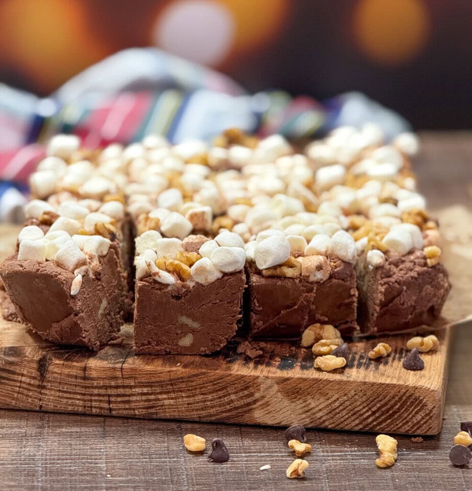 Sliced chocolate fudge topped with mini marshmallows and chopped nuts on a wooden board, with a colorful cloth and blurred lights in the background.
