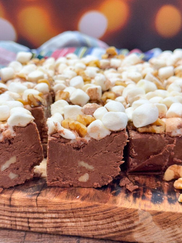 Close-up of chocolate fudge squares topped with mini marshmallows and chopped walnuts, arranged on a wooden surface. The background is softly blurred with warm lights.