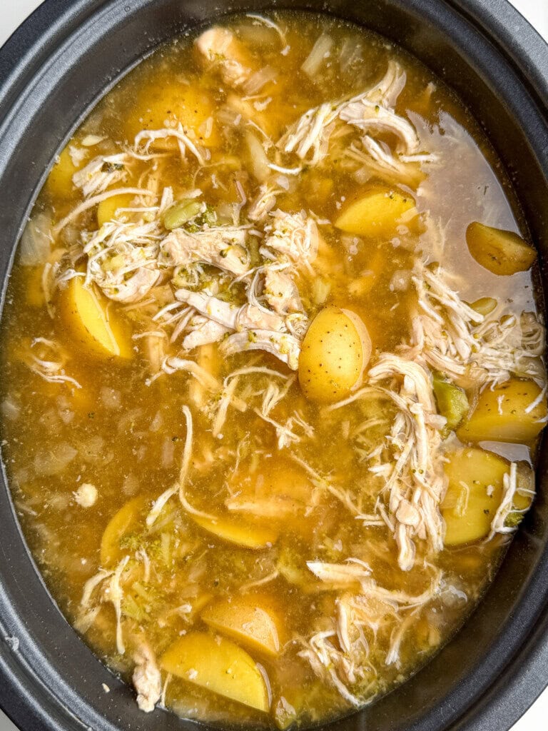 A close-up of a pot filled with shredded chicken, yellow potato pieces, and a greenish broth with herbs and spices.