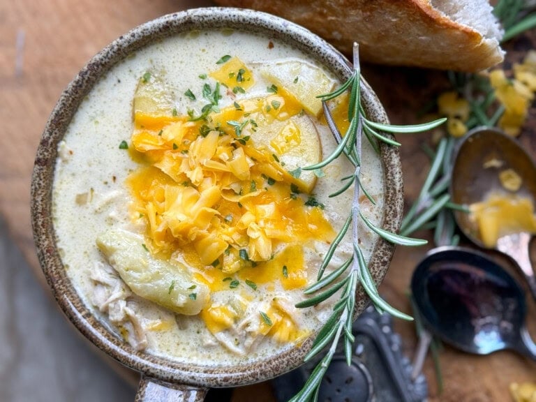 A bowl of creamy soup topped with shredded cheddar cheese and fresh herbs, garnished with a rosemary sprig. Potato pieces are visible in the soup, with a piece of bread and spoons in the background.