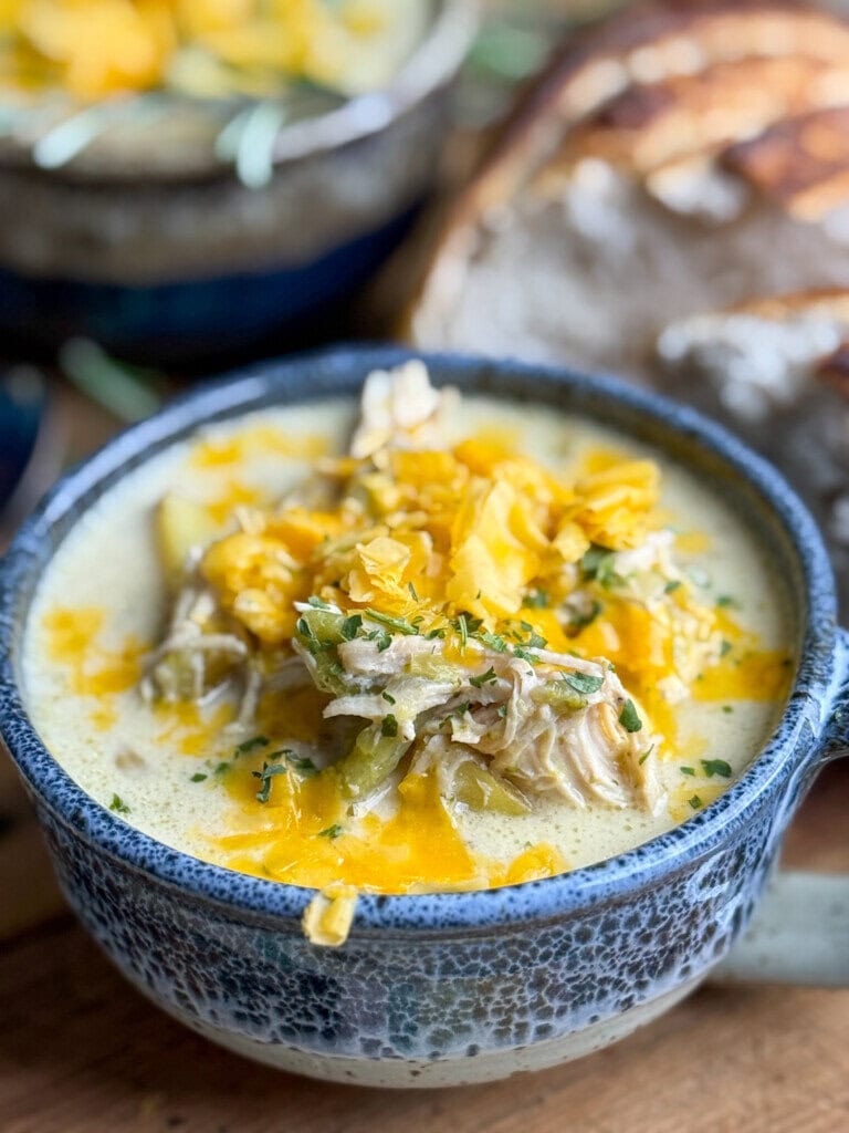 A rustic blue ceramic bowl filled with creamy chicken soup, topped with shredded cheese and herbs. Slices of crusty bread are in the background, suggesting a cozy meal setting.