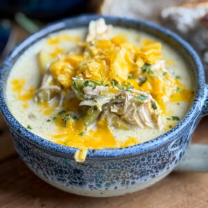 A close-up of a blue ceramic bowl filled with creamy chicken soup, shredded chicken, potatoes, chopped herbs, and topped with shredded cheddar cheese, with a slice of bread in the background.