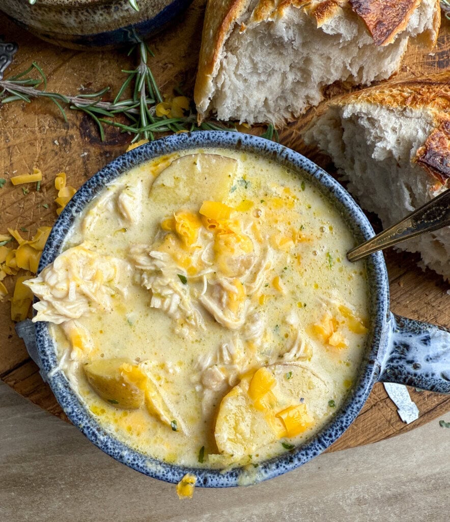 A bowl of creamy chicken and potato soup garnished with shredded cheese sits on a wooden surface, next to slices of rustic bread and fresh herbs.
