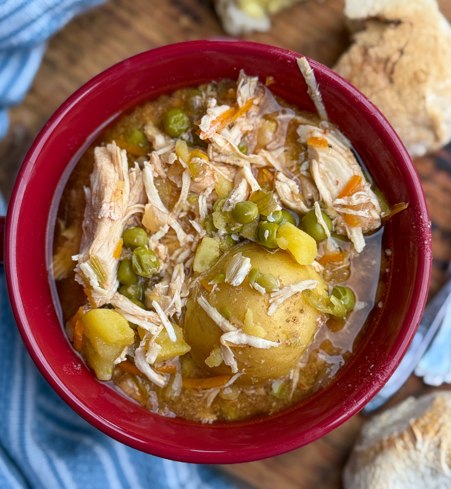 A bowl of chicken stew with shredded chicken, potatoes, peas, carrots, and broth, served in a red bowl. Pieces of crusty bread and a blue cloth are nearby on a wooden surface.