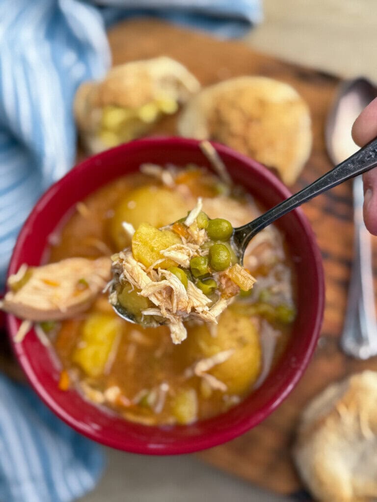 A spoonful of chicken soup with peas, potatoes, and shredded chicken is held above a red bowl, with bread rolls and a blue-striped cloth in the background.