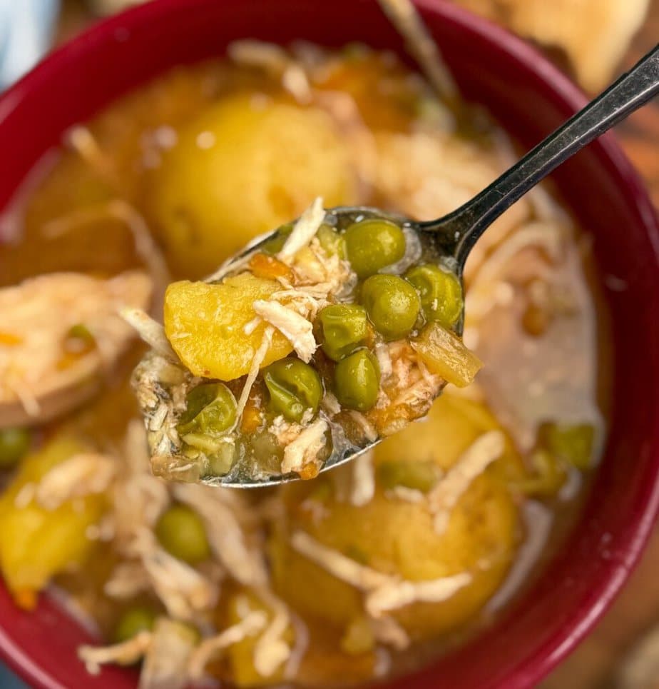 A close-up of a spoonful of soup with shredded chicken, peas, and diced potatoes held over a red bowl filled with the same hearty soup.