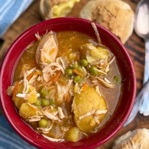 A red bowl filled with chicken stew containing shredded chicken, potatoes, peas, and carrots, placed next to two bread rolls on a wooden board with a striped blue napkin and silver utensils.
