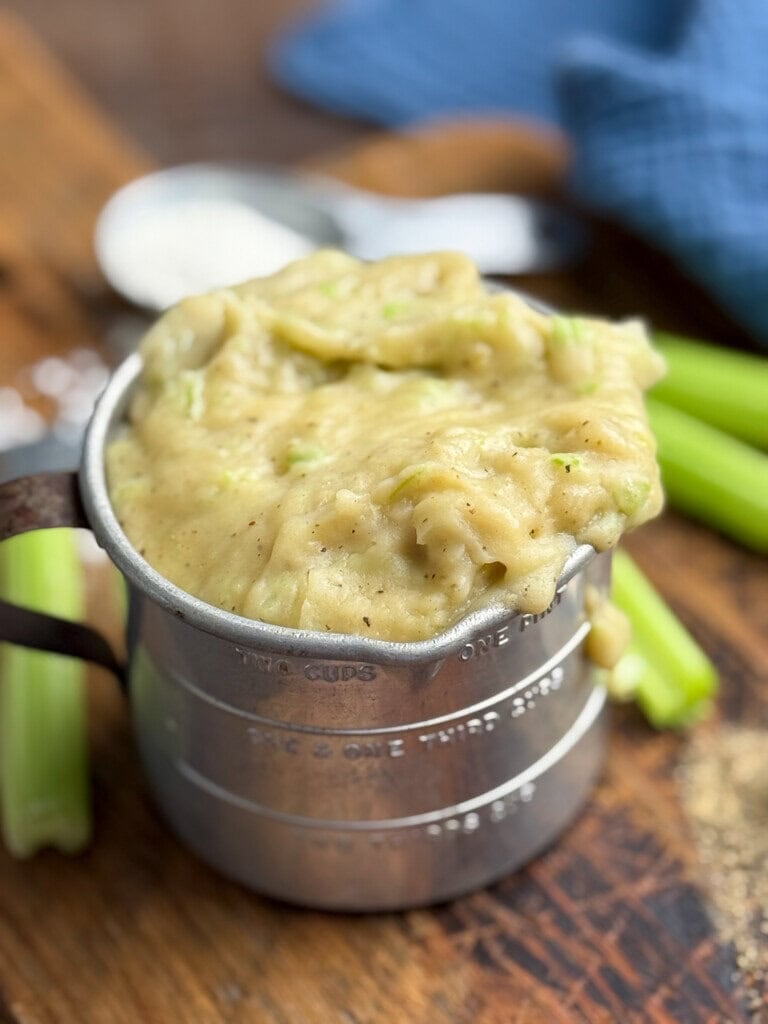 A metal measuring cup filled with thick, creamy celery soup sits on a wooden surface. Chopped celery stalks and a blue cloth are visible in the background. The soup is overflowing slightly from the cup.