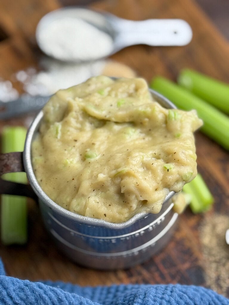 A metal measuring cup filled with a creamy mixture containing small green celery pieces sits on a wooden surface, with celery stalks and a scoop of flour in the background.