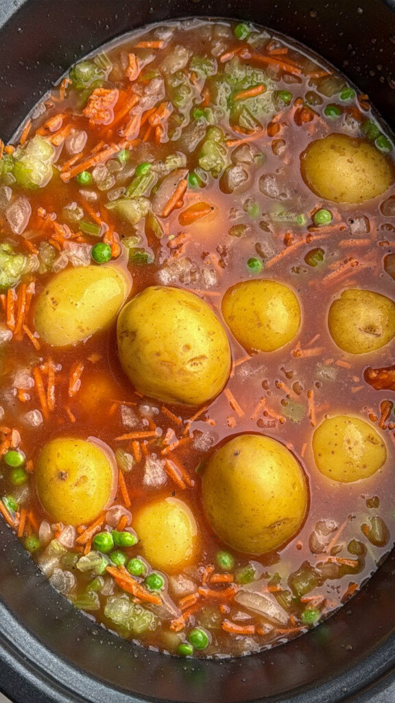 A close-up of a pot filled with vegetable stew, featuring whole baby potatoes, green peas, diced green bell peppers, shredded carrots, onions, and a red-brown broth.