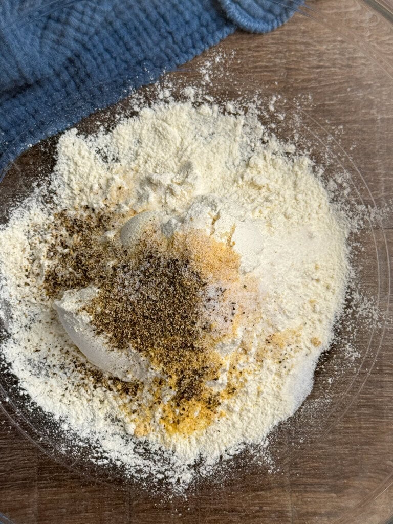 A clear bowl on a wooden surface contains a mixture of flour, black pepper, salt, and other powdered spices next to a blue textured cloth.