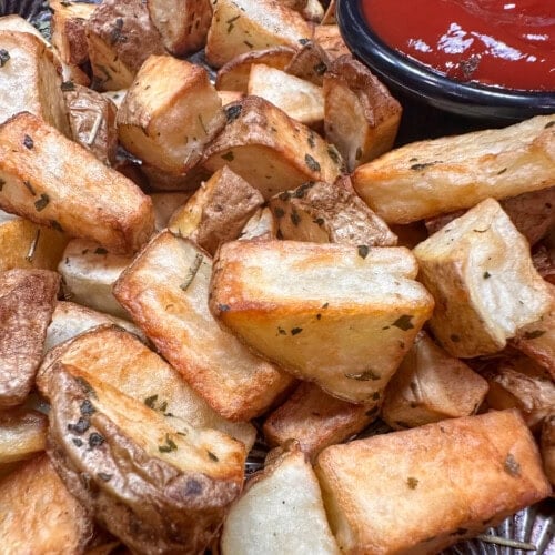 A close-up of crispy roasted potato wedges garnished with herbs, served next to a small black bowl filled with ketchup.
