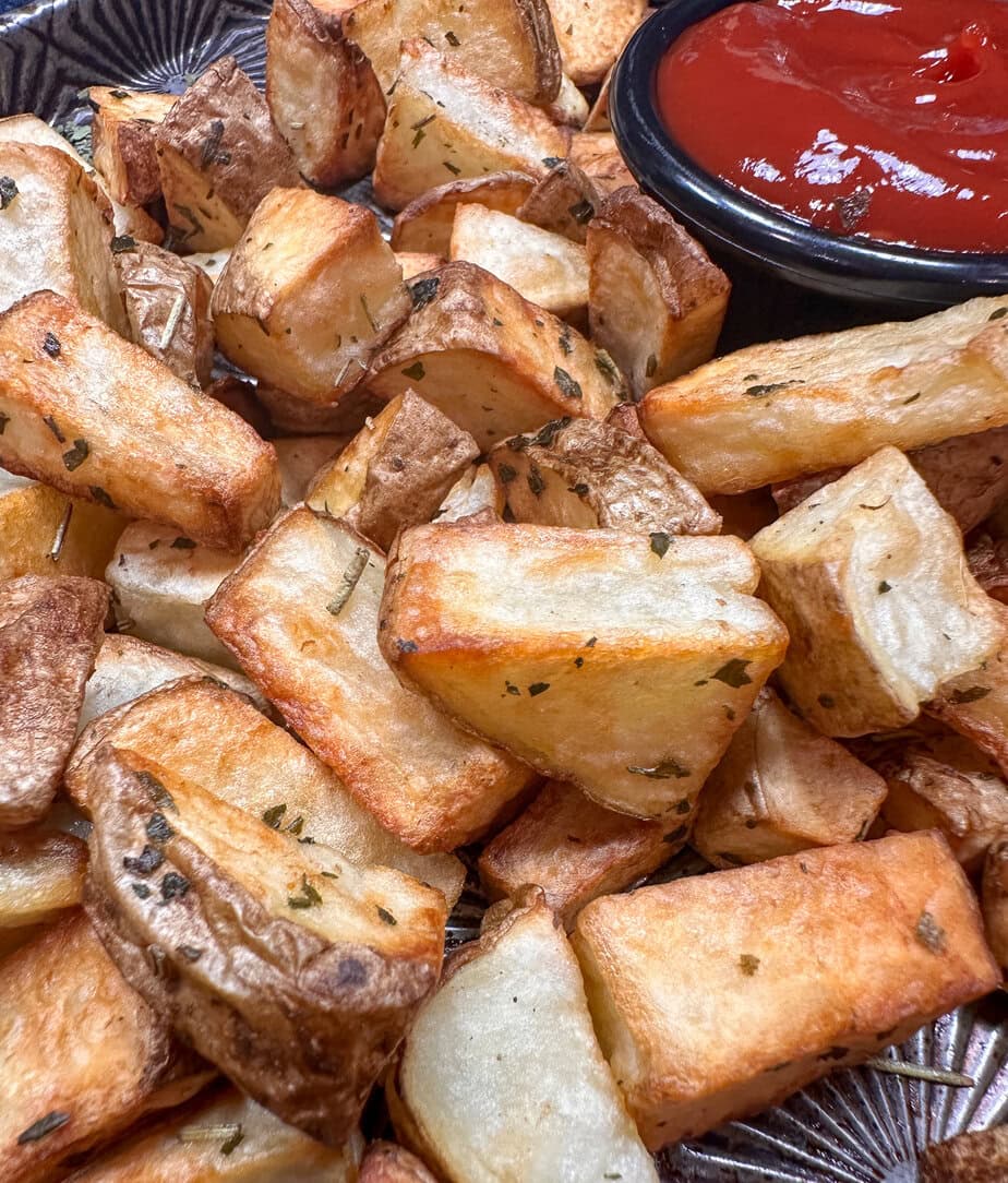 A close-up of crispy roasted potato wedges garnished with herbs, served next to a small black bowl filled with ketchup.