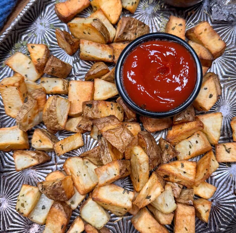 A tray of roasted potato cubes seasoned with herbs surrounds a small black bowl filled with ketchup.