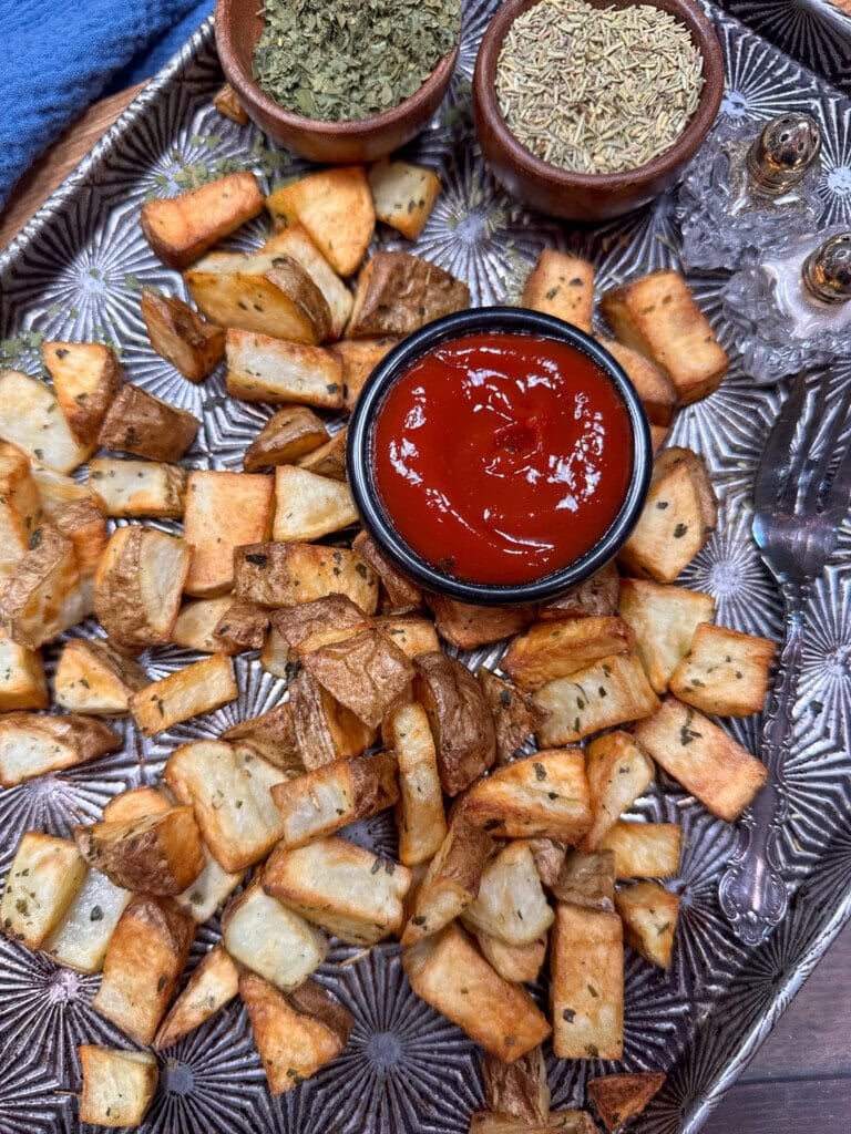 A tray of roasted potato cubes seasoned with herbs, served with a bowl of ketchup in the center. Small bowls of dried herbs and a pair of salt and pepper shakers are also on the tray.