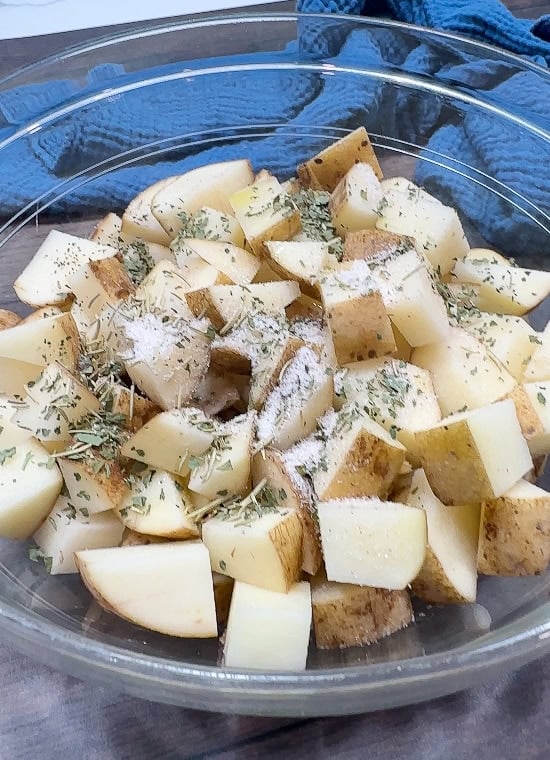 Clear glass bowl filled with chopped potatoes, topped with dried herbs and seasoning, sitting on a wooden surface with a blue cloth in the background.