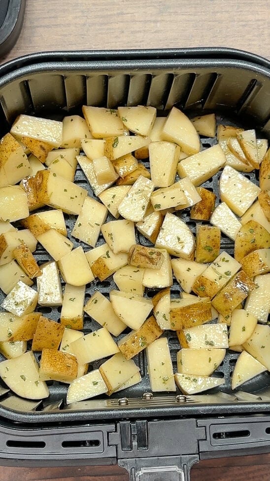 Cubed potatoes with herbs and seasoning arranged in a single layer inside an air fryer basket, ready to be cooked.