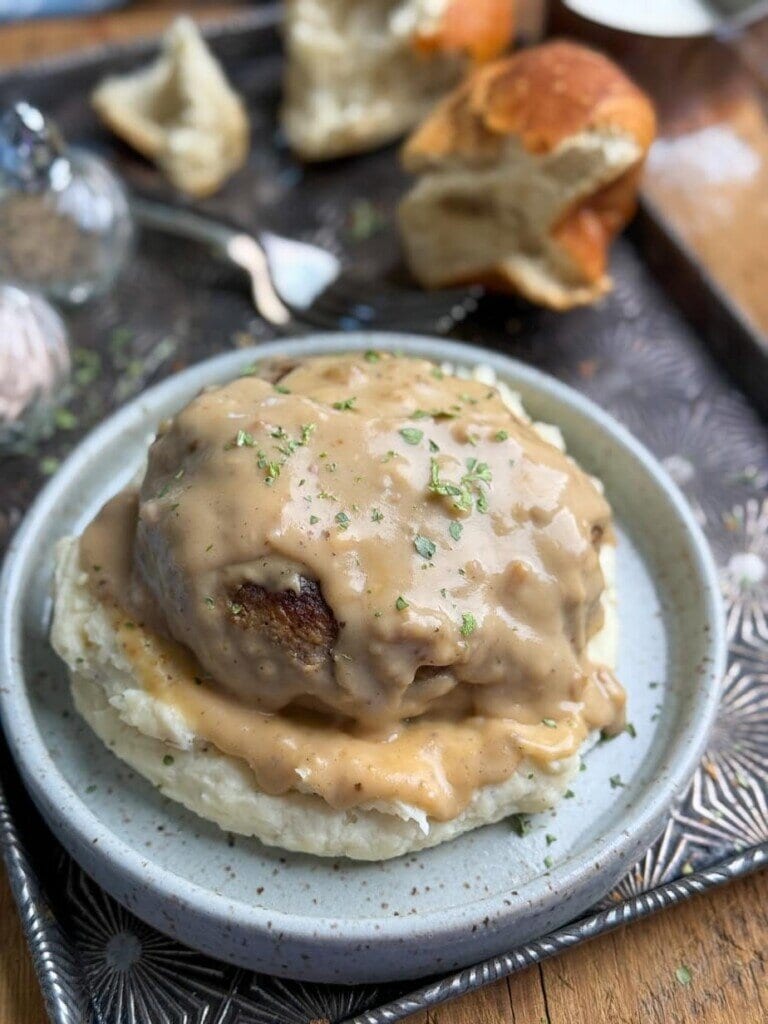 A plate with mashed potatoes topped with a large meatball and creamy brown gravy, garnished with chopped herbs. In the background, pieces of bread and a salt shaker rest on a metal tray.