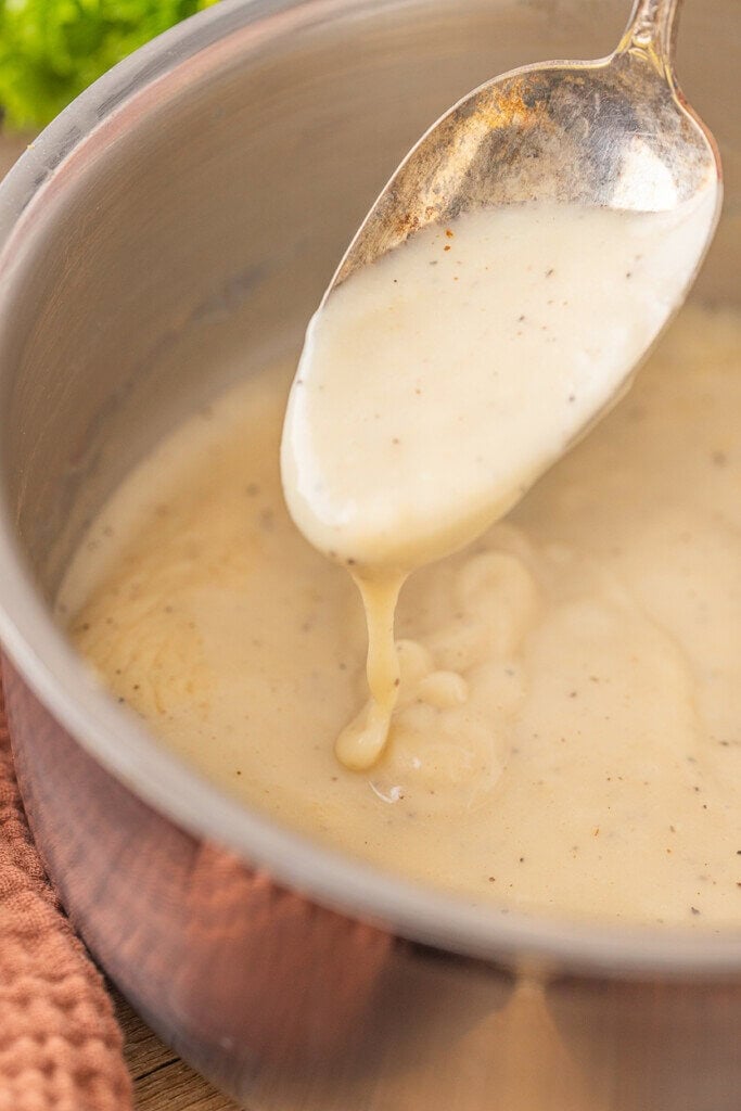 A spoon lifting creamy condensed chicken soup from a saucepan, with the sauce dripping back into the pan. The sauce appears smooth and speckled with black pepper. A brown towel is partially visible beside the pan.