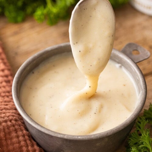 A spoon is lifting creamy condensed chicken soup out of a ceramic bowl on a wooden surface, with fresh parsley and a textured brown cloth nearby.