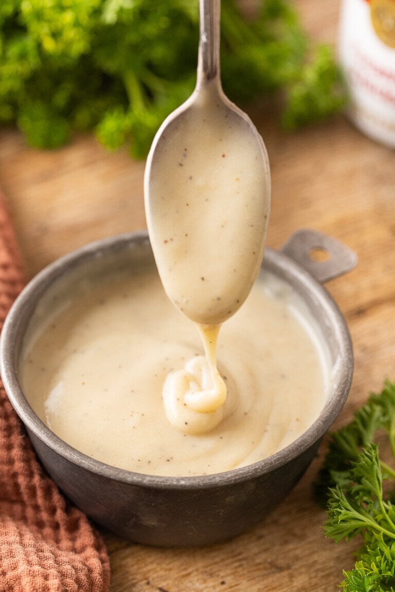 A spoon drizzles creamy white condensed chicken soup with black pepper specks into a ceramic bowl, set on a wooden surface with fresh parsley and a brown cloth nearby.