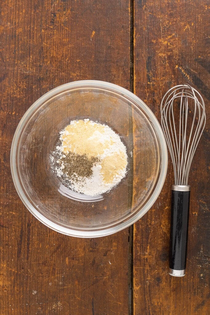 A glass bowl with spices and flour, including black pepper and garlic powder, sits on a wooden table next to a metal whisk with a black handle.
