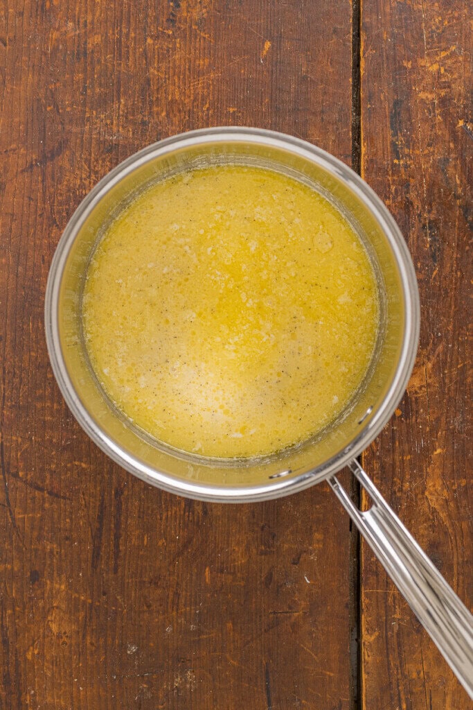 A metal saucepan filled with melted butter sits on a rustic wooden surface, viewed from above. The mixture is yellow and slightly frothy around the edges.