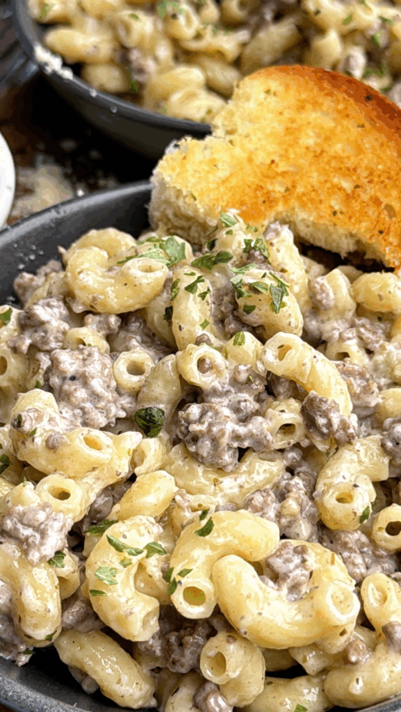 A close-up of creamy macaroni pasta mixed with ground beef and garnished with chopped herbs, served with a slice of toasted garlic bread on the side.