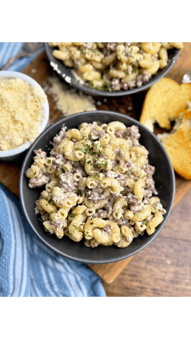 A black bowl filled with creamy macaroni and ground beef, garnished with herbs, sits on a wooden board next to a slice of garlic bread and a small bowl of grated cheese. Another bowl of pasta is visible in the background.
