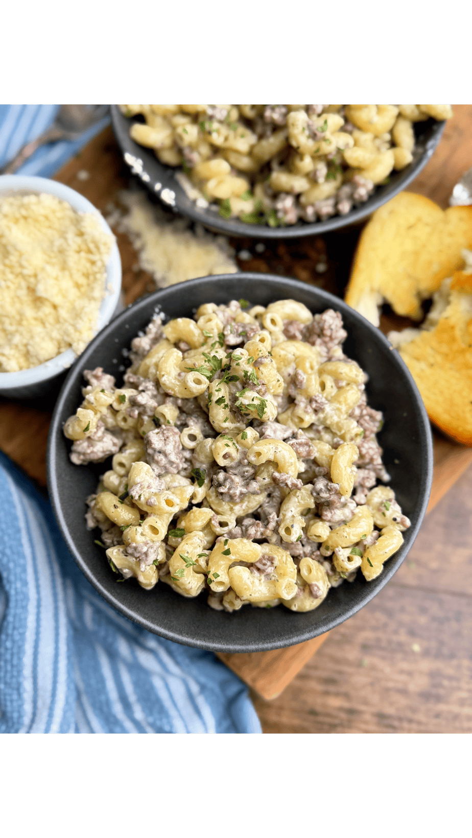 A black bowl filled with creamy macaroni and ground beef, garnished with herbs, sits on a wooden board next to a slice of garlic bread and a small bowl of grated cheese. Another bowl of pasta is visible in the background.