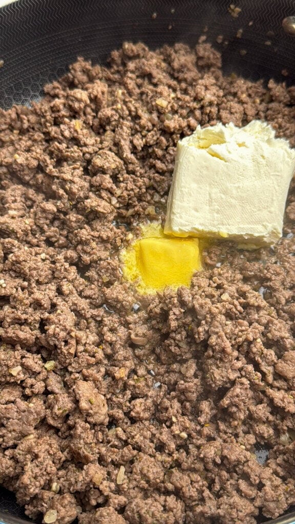 Ground beef cooking in a pan with a block of cream cheese and a pat of butter placed on top, ready to be mixed in.