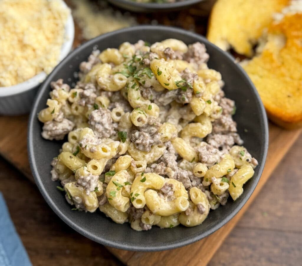 A black bowl filled with creamy macaroni and ground beef, garnished with chopped herbs, sits on a wooden surface next to a slice of buttered bread and a dish of shredded cheese.