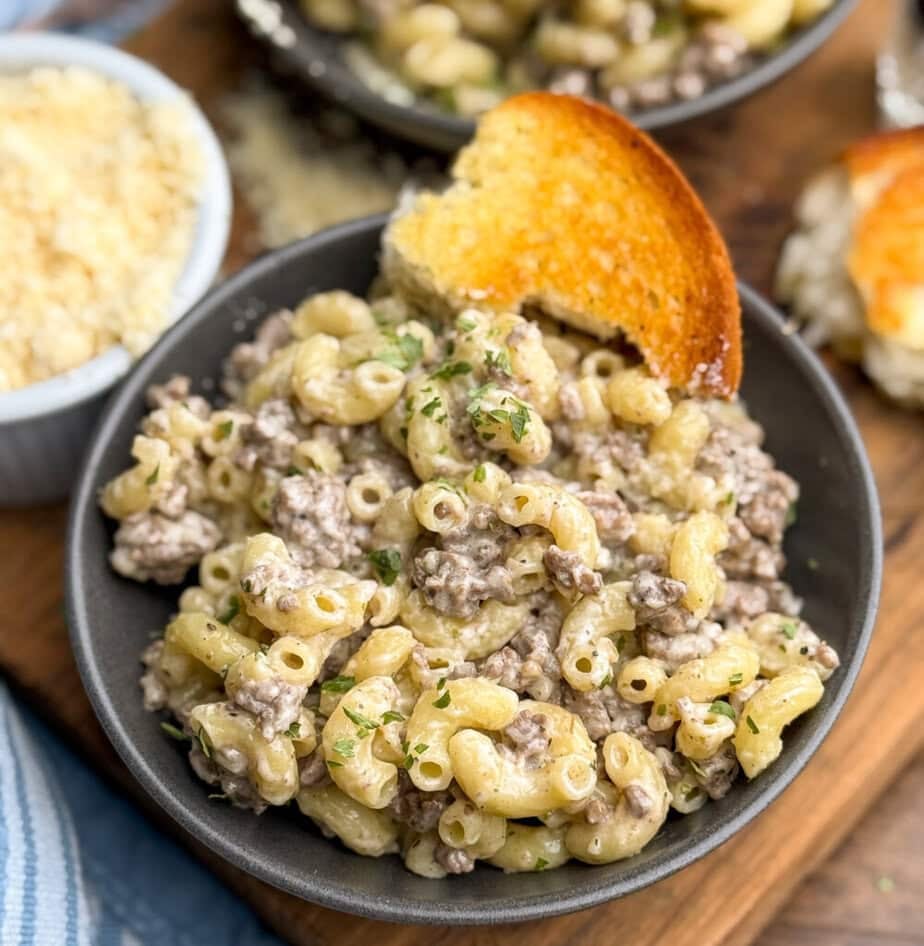 A bowl of creamy macaroni with ground beef, garnished with herbs, and served with a slice of toasted bread. A dish of grated cheese is in the background on a wooden surface.