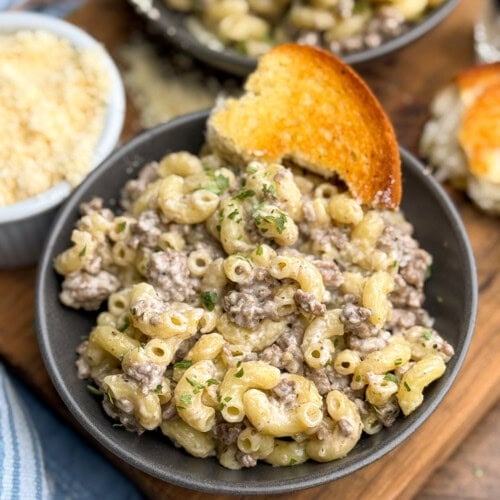 A bowl of creamy macaroni with ground beef, garnished with herbs, and served with a slice of toasted bread. A dish of grated cheese is in the background on a wooden surface.