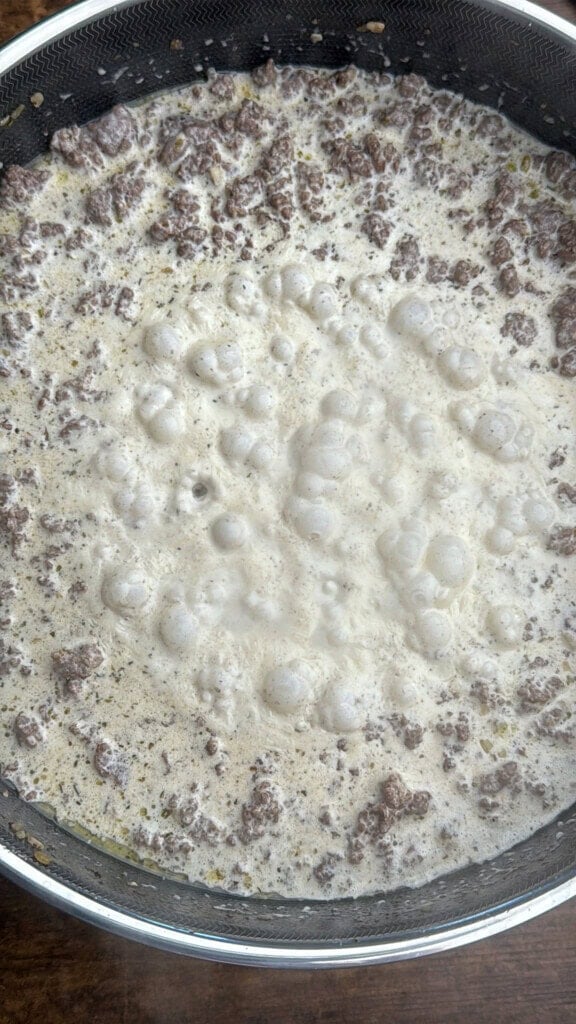 Ground beef simmering in a skillet with a creamy white sauce, creating bubbles on the surface as it cooks.