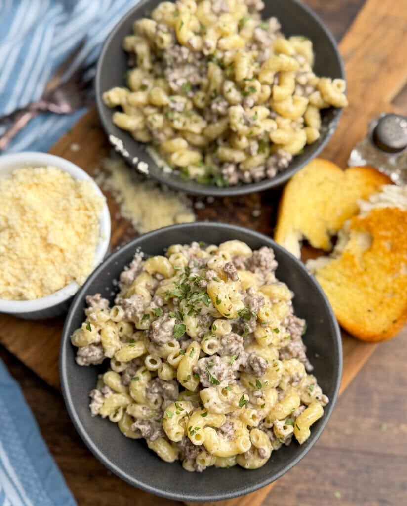 Two black bowls filled with creamy macaroni and ground beef, garnished with herbs, sit on a wooden board. Nearby are slices of toasted bread and a small bowl of grated cheese. A blue-striped cloth is partially visible.