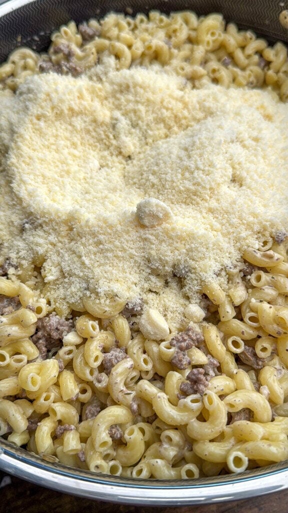 A close-up of cooked elbow macaroni mixed with ground beef, topped with a generous layer of grated parmesan cheese in a large pot.